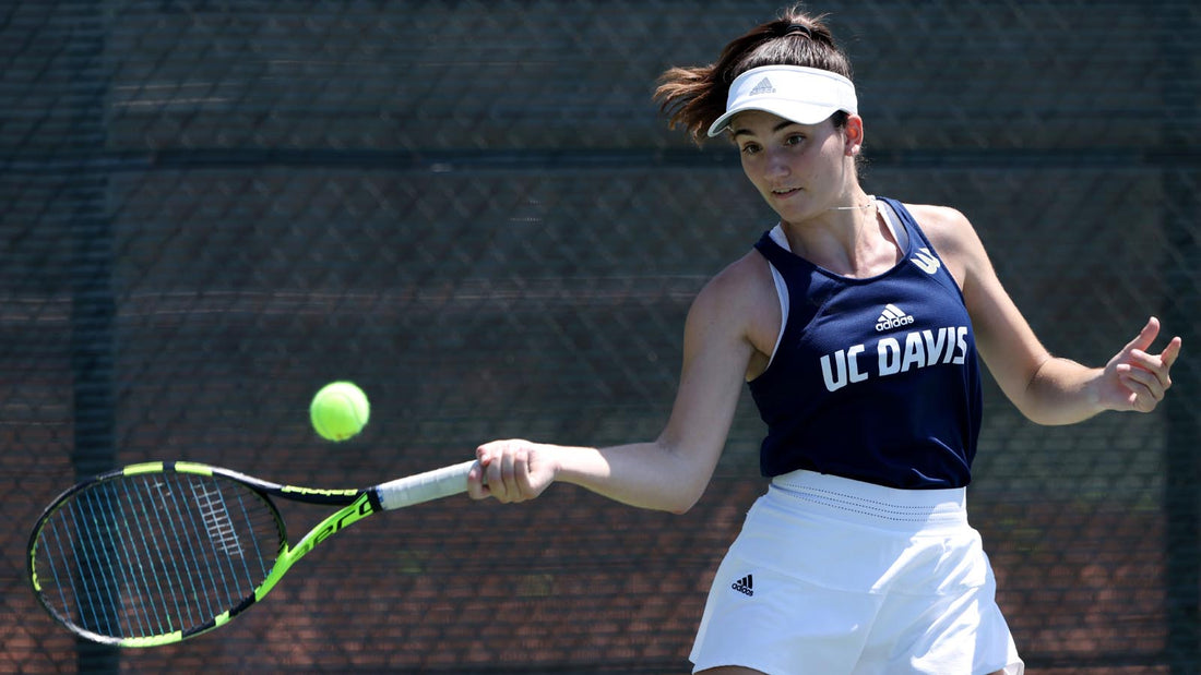 Female college tennis player hits a forehand while wearing blue UC Davis shirt with white lettering and white Adidas skirt