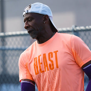 Man in orange shirt with 'BEAST' text holding a paddle on a tennis court.