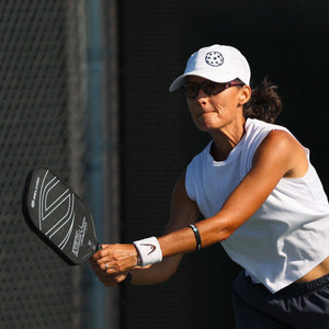 A woman plays pickleball, hitting the ball mid-air with intensity. She wears a white cap, sleeveless top, and dark shorts on a sunny court.