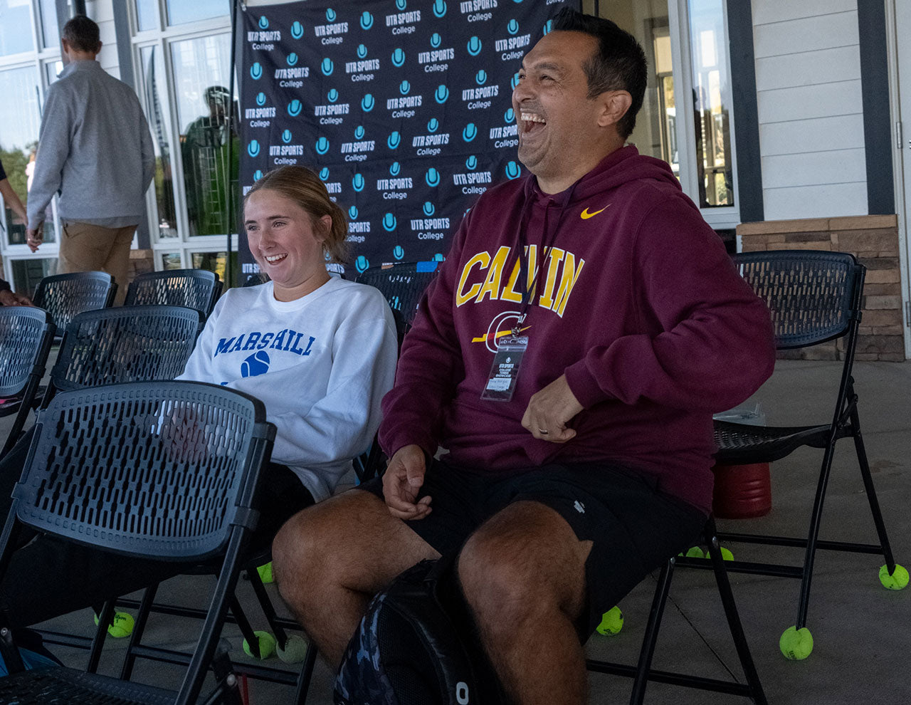 Two people sitting in black folding chairs in front of a UTR Sports backdrop. The person on the right wears a maroon hoodie with yellow 'CALVIN' lettering and a Nike swoosh, while the person on the left wears a white sweatshirt with 'Marshall' and a blue btennisl graphic.